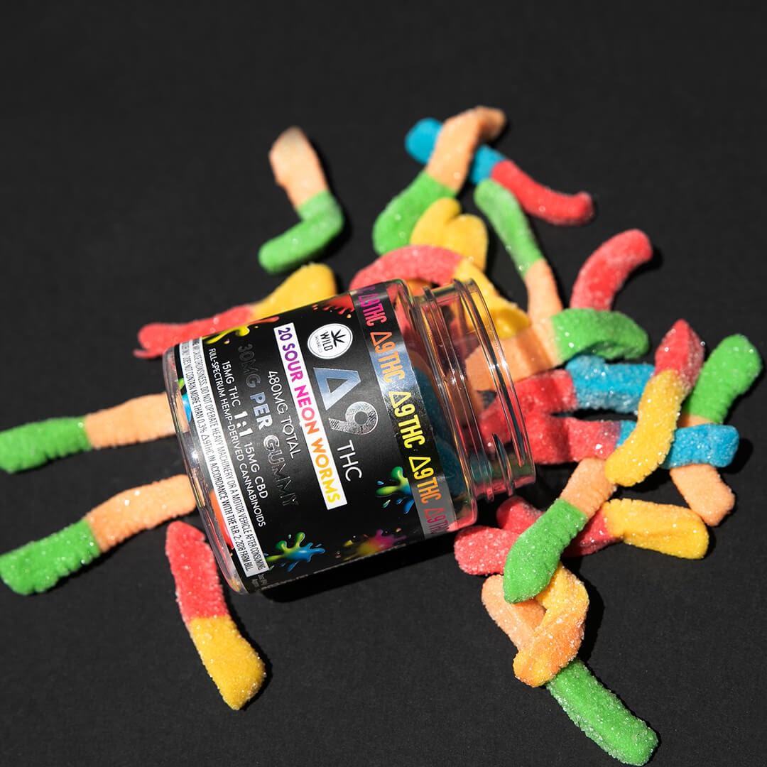 A tipped jar labeled "Delta Astro Acid Trip" with colorful sugar-coated gummy worms scattered on a black background, evoking the vibrant essence of an HHC-R experience.