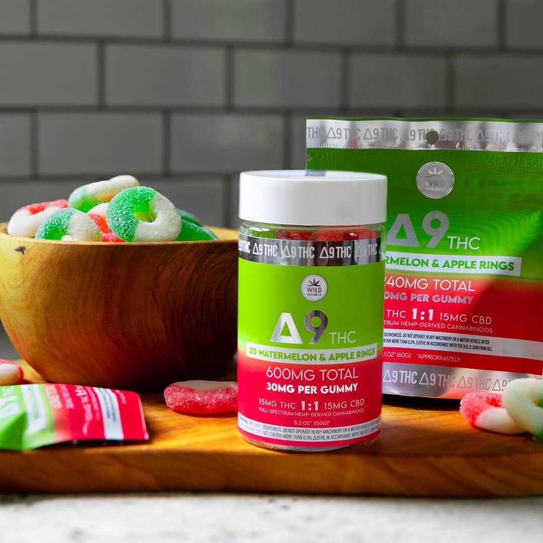 Jars and a bag of Delta 9 THC watermelon and apple rings sit on a wooden board, with a bowl of gummies beside them. The packaging displays THC, CBD, and THCa content for informed indulgence.