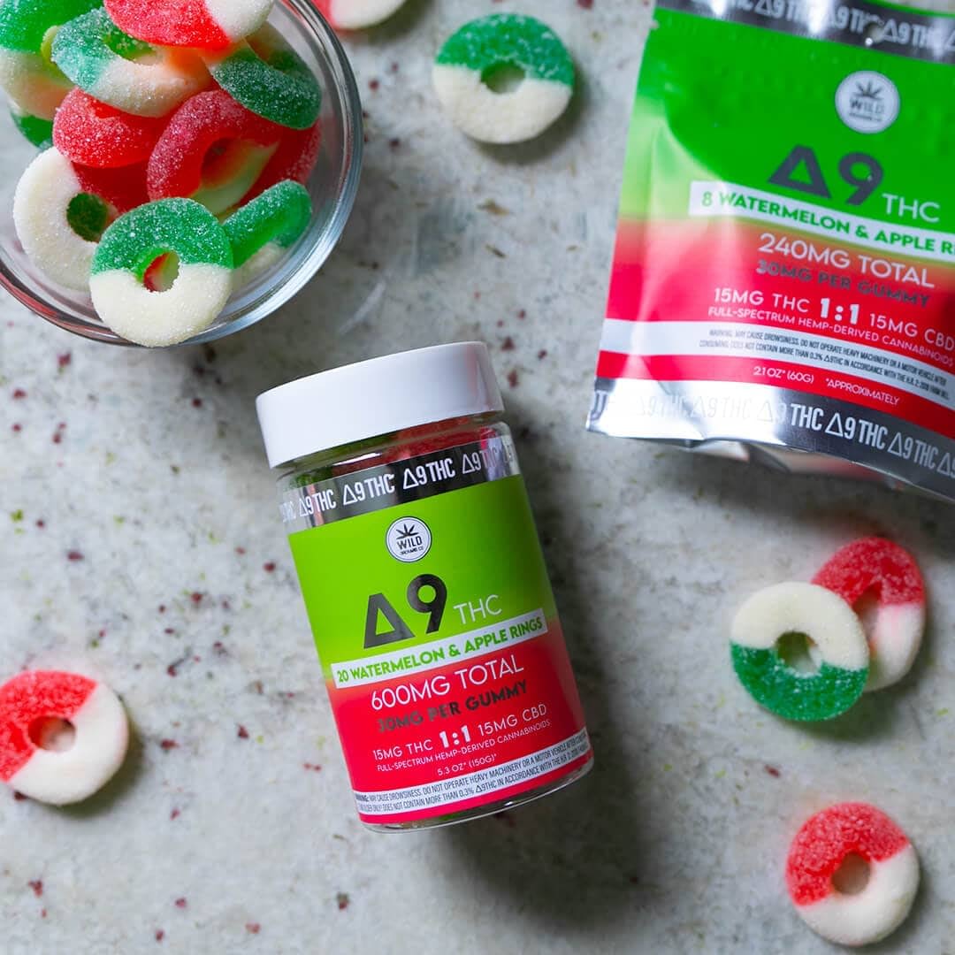 A jar of D9 THC gummy rings sits beside a bowl of similar edibles and an open package on a speckled surface.