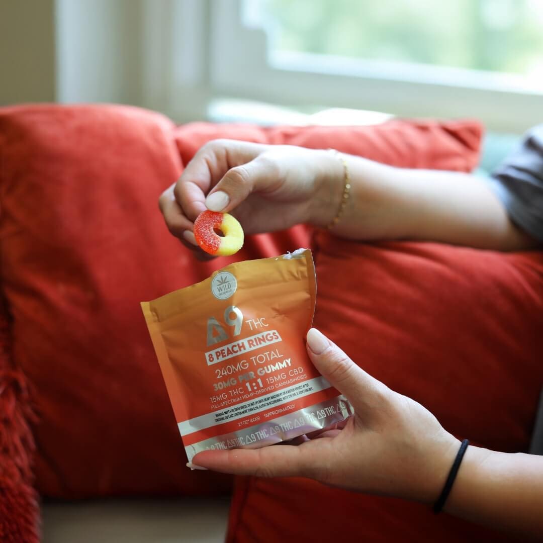 A person holds a peach ring gummy above an open package labeled “Delta 9 Peach Rings” with dosage information, sitting on an orange couch by a window.