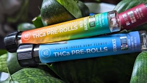 Two pre-roll containers labeled "THCa PRE-ROLLS" resting on a pile of green produce. The labels read "Jolly Rancher" and "Cookies," with "Indica" and "Sativa" indicated respectively.