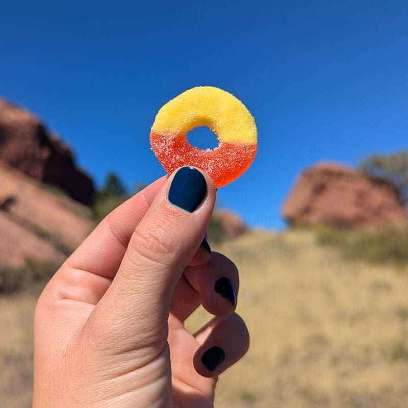 A hand with dark blue nail polish holds a vibrant yellow and orange Delta 9 Peach Ring gummy against a clear blue sky, with a blurred rocky landscape stretching out behind it.